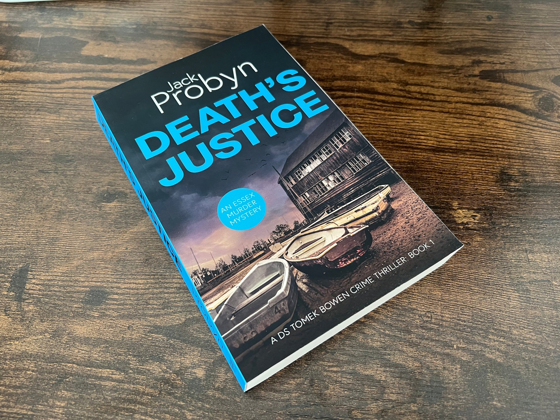 Book cover of Death's Justice, an Essex Murder Mystery by Jack Probyn. A gritty crime thriller featuring a weathered wooden boathouse on a muddy Essex shoreline at dusk. Book 1 in the DS Tomek Bowen series, perfect for fans of JD Kirk and JM Dalgliesh
